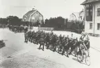 En cyklist och män till häst i uniform m/1910 leder marscherande trupp i tre grupper.