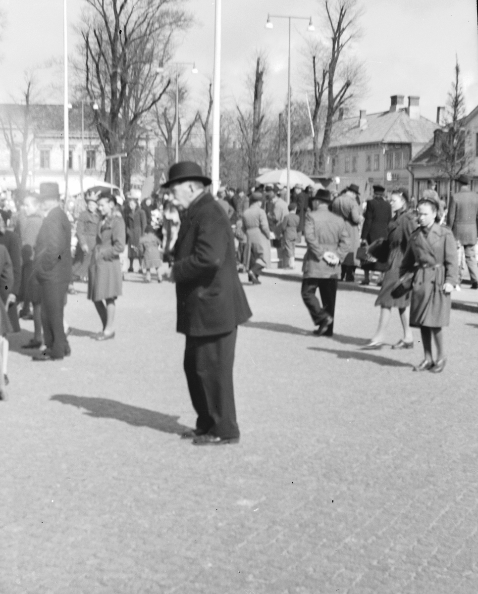 Miljöbild från Varbergs torg med en äldre man i fokus. Han och flera andra verkar betrakta något som händer till vänster ur bild. I bakgrunden en folksamling och till höger ses kv Apoteket.