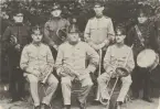 Skaraborgs flygflottilj F7, flygvapensymbolen med texten Första flygeskadern.Karlsborgs fästning. Artilleriets musikkår, sju män poserar i trädgården med sina instrument. Uniform m/1910.