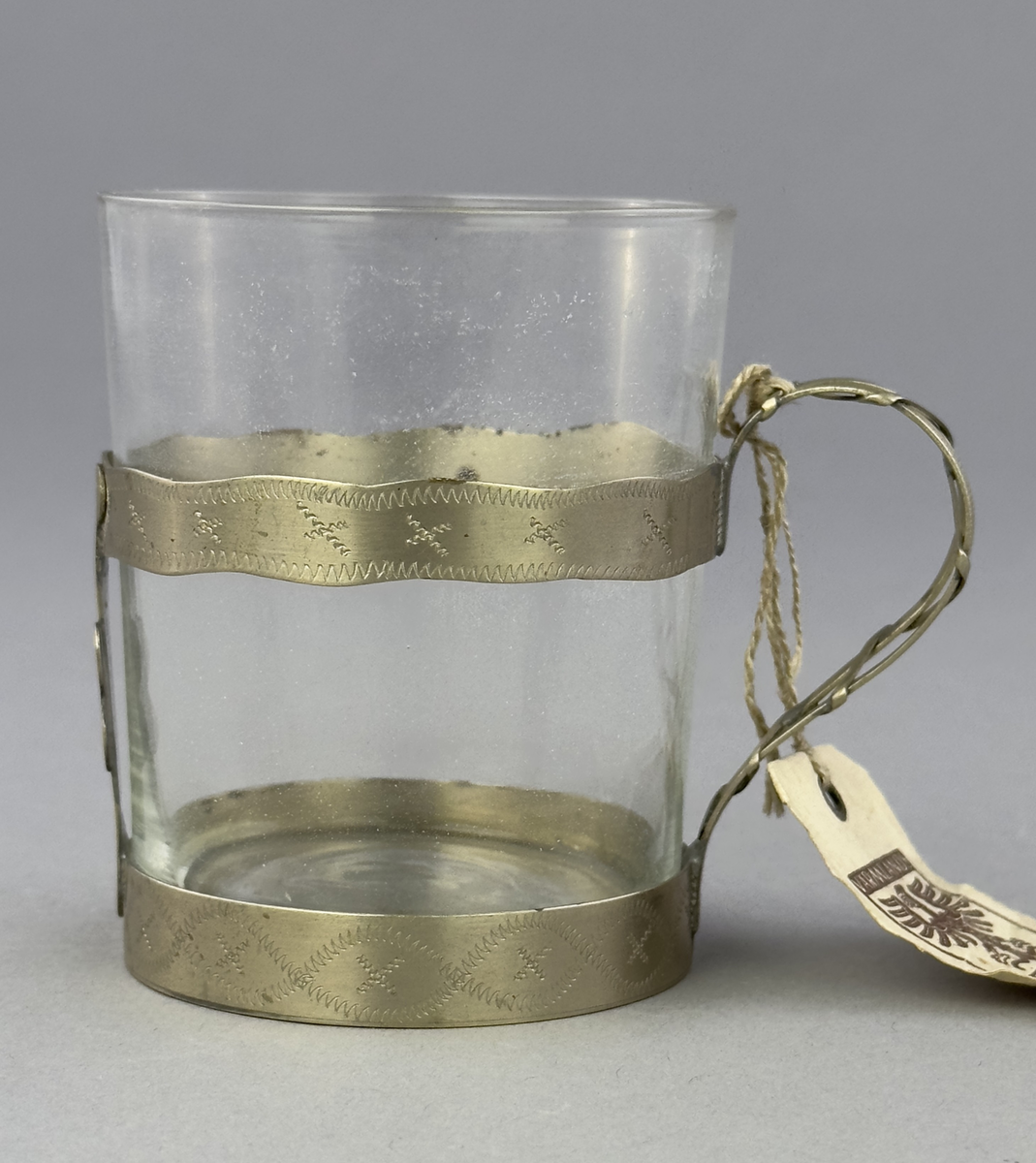 Temugg av glas och tenn. Tennhållaren är dekorerad med ristat mönster.