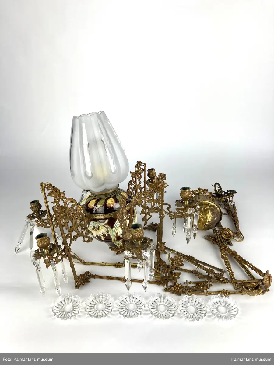 KLM 45619:390. Lampa, taklampa, fotogenlampa. Ombyggd fotogenlampa med stomme av bronserad gjutmetall samt keramik. Mittendelen är av glaserad keramik/porslin med gula blommor på stjälk med gröna blad. Bottenfärg i rödbrun nyans. Övre delen  är avtagbar med fastsatt glasbehållare undertill för fotogen. Lampan är ombyggd med dragen el och hållare för glödlampa. Sammanlagt 6 armar med hållare för stearinljus. Ljusarmarna är i snirklig jugendstil och är förgyllda. Ytterst på vardera arm hänger prismor av typen fotogenprisma. 6 tillhörande ljusmanschetter i genomskinligt glas. Glaskupa som sitter överst på stommen, i frostat vitt glas med sparade ytor som bildar mönster. Takkronan hänger i avlånga metalldelar som är ihopsatta med metallringar och fungerar på så vis som en kedja. Alla dekorerade med jugendmönster. Överst en hissanordning.