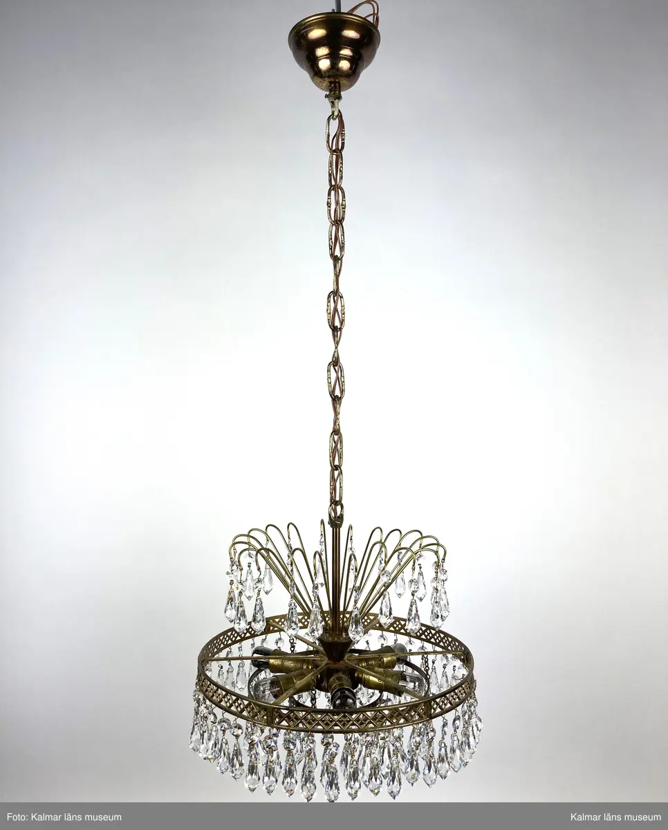 KLM45619:391. Lampa, taklampa, kristallkrona. Av mässing med tre ringar av olika diametrar som hänger på olika höjd med den minsta längst ner. På ringarna är prismor fastsatta, överst en förprisma av sorten oktagon och undertill fäst i denna en prisma av sorten pigg. Överst 20 stycken böjda mässingspinnar med likadan sammansättning av prismor som på ringarna beskrivna ovan. Elektrifierad med plats för 6 glödlampor.