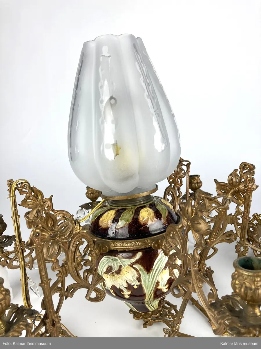KLM 45619:390. Lampa, taklampa, fotogenlampa. Ombyggd fotogenlampa med stomme av bronserad gjutmetall samt keramik. Mittendelen är av glaserad keramik/porslin med gula blommor på stjälk med gröna blad. Bottenfärg i rödbrun nyans. Övre delen  är avtagbar med fastsatt glasbehållare undertill för fotogen. Lampan är ombyggd med dragen el och hållare för glödlampa. Sammanlagt 6 armar med hållare för stearinljus. Ljusarmarna är i snirklig jugendstil och är förgyllda. Ytterst på vardera arm hänger prismor av typen fotogenprisma. 6 tillhörande ljusmanschetter i genomskinligt glas. Glaskupa som sitter överst på stommen, i frostat vitt glas med sparade ytor som bildar mönster. Takkronan hänger i avlånga metalldelar som är ihopsatta med metallringar och fungerar på så vis som en kedja. Alla dekorerade med jugendmönster. Överst en hissanordning.
