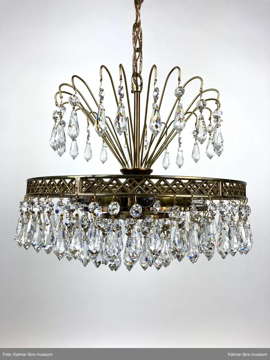 KLM45619:391. Lampa, taklampa, kristallkrona. Av mässing med tre ringar av olika diametrar som hänger på olika höjd med den minsta längst ner. På ringarna är prismor fastsatta, överst en förprisma av sorten oktagon och undertill fäst i denna en prisma av sorten pigg. Överst 20 stycken böjda mässingspinnar med likadan sammansättning av prismor som på ringarna beskrivna ovan. Elektrifierad med plats för 6 glödlampor.