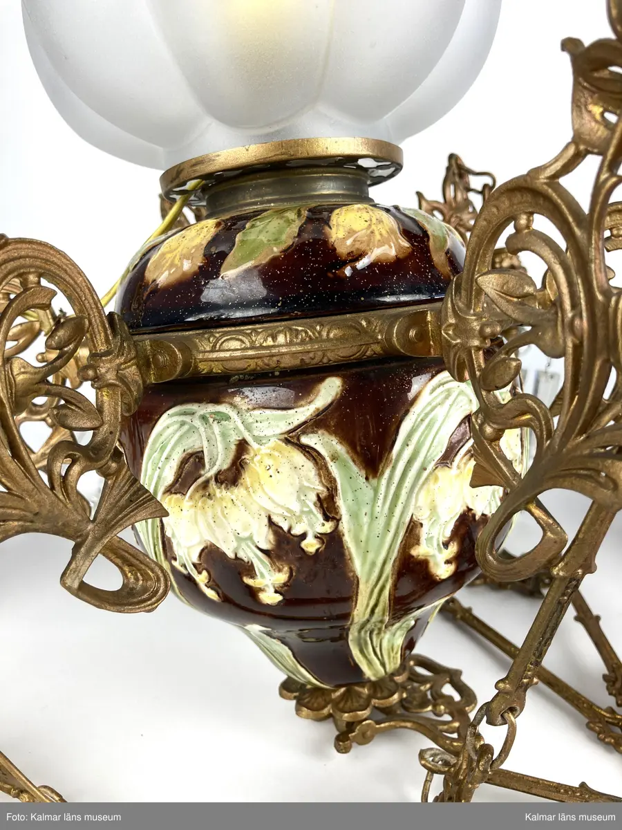 KLM 45619:390. Lampa, taklampa, fotogenlampa. Ombyggd fotogenlampa med stomme av bronserad gjutmetall samt keramik. Mittendelen är av glaserad keramik/porslin med gula blommor på stjälk med gröna blad. Bottenfärg i rödbrun nyans. Övre delen  är avtagbar med fastsatt glasbehållare undertill för fotogen. Lampan är ombyggd med dragen el och hållare för glödlampa. Sammanlagt 6 armar med hållare för stearinljus. Ljusarmarna är i snirklig jugendstil och är förgyllda. Ytterst på vardera arm hänger prismor av typen fotogenprisma. 6 tillhörande ljusmanschetter i genomskinligt glas. Glaskupa som sitter överst på stommen, i frostat vitt glas med sparade ytor som bildar mönster. Takkronan hänger i avlånga metalldelar som är ihopsatta med metallringar och fungerar på så vis som en kedja. Alla dekorerade med jugendmönster. Överst en hissanordning.