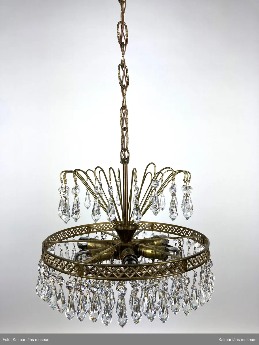 KLM45619:391. Lampa, taklampa, kristallkrona. Av mässing med tre ringar av olika diametrar som hänger på olika höjd med den minsta längst ner. På ringarna är prismor fastsatta, överst en förprisma av sorten oktagon och undertill fäst i denna en prisma av sorten pigg. Överst 20 stycken böjda mässingspinnar med likadan sammansättning av prismor som på ringarna beskrivna ovan. Elektrifierad med plats för 6 glödlampor.