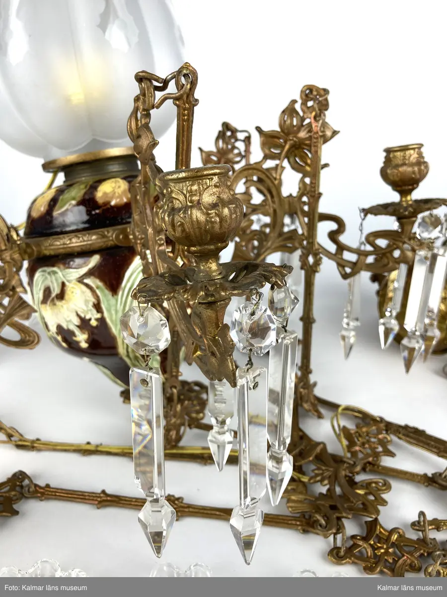 KLM 45619:390. Lampa, taklampa, fotogenlampa. Ombyggd fotogenlampa med stomme av bronserad gjutmetall samt keramik. Mittendelen är av glaserad keramik/porslin med gula blommor på stjälk med gröna blad. Bottenfärg i rödbrun nyans. Övre delen  är avtagbar med fastsatt glasbehållare undertill för fotogen. Lampan är ombyggd med dragen el och hållare för glödlampa. Sammanlagt 6 armar med hållare för stearinljus. Ljusarmarna är i snirklig jugendstil och är förgyllda. Ytterst på vardera arm hänger prismor av typen fotogenprisma. 6 tillhörande ljusmanschetter i genomskinligt glas. Glaskupa som sitter överst på stommen, i frostat vitt glas med sparade ytor som bildar mönster. Takkronan hänger i avlånga metalldelar som är ihopsatta med metallringar och fungerar på så vis som en kedja. Alla dekorerade med jugendmönster. Överst en hissanordning.