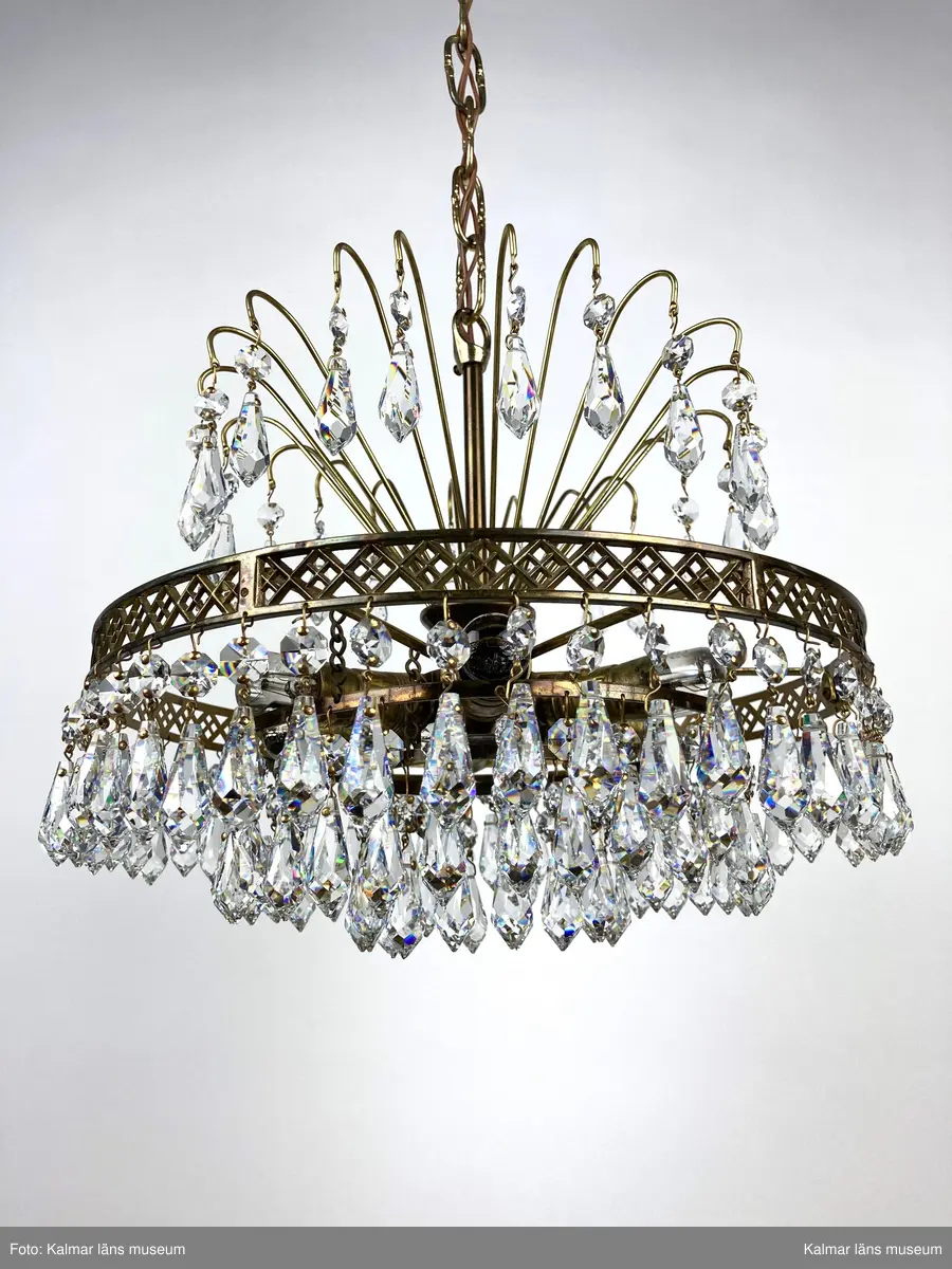 KLM45619:391. Lampa, taklampa, kristallkrona. Av mässing med tre ringar av olika diametrar som hänger på olika höjd med den minsta längst ner. På ringarna är prismor fastsatta, överst en förprisma av sorten oktagon och undertill fäst i denna en prisma av sorten pigg. Överst 20 stycken böjda mässingspinnar med likadan sammansättning av prismor som på ringarna beskrivna ovan. Elektrifierad med plats för 6 glödlampor.