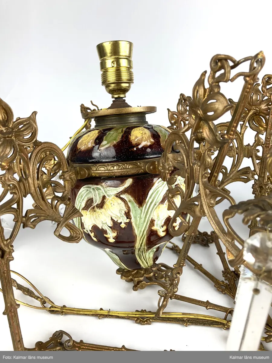 KLM 45619:390. Lampa, taklampa, fotogenlampa. Ombyggd fotogenlampa med stomme av bronserad gjutmetall samt keramik. Mittendelen är av glaserad keramik/porslin med gula blommor på stjälk med gröna blad. Bottenfärg i rödbrun nyans. Övre delen  är avtagbar med fastsatt glasbehållare undertill för fotogen. Lampan är ombyggd med dragen el och hållare för glödlampa. Sammanlagt 6 armar med hållare för stearinljus. Ljusarmarna är i snirklig jugendstil och är förgyllda. Ytterst på vardera arm hänger prismor av typen fotogenprisma. 6 tillhörande ljusmanschetter i genomskinligt glas. Glaskupa som sitter överst på stommen, i frostat vitt glas med sparade ytor som bildar mönster. Takkronan hänger i avlånga metalldelar som är ihopsatta med metallringar och fungerar på så vis som en kedja. Alla dekorerade med jugendmönster. Överst en hissanordning.