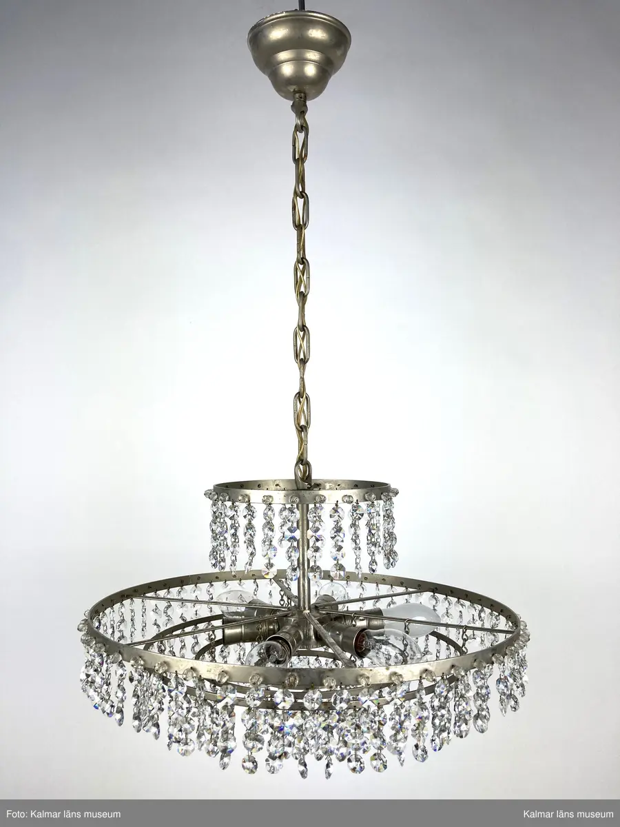 KLM 45619:392. Lampa, taklampa, kristallkrona. Av vit metall med fyra ringar av olika diametrar som hänger på olika höjd. På ringarna är prismor av sorten koppen fastsatta. På de två största ringarna sitter även pärlor fästa direkt i ringen. Eleiktrifierad med plats för 6 glödlampor.
