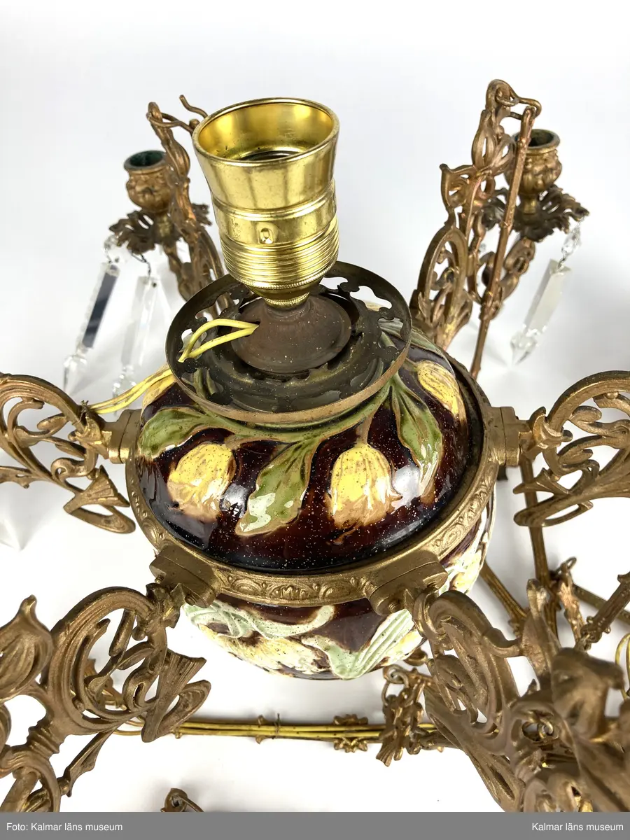 KLM 45619:390. Lampa, taklampa, fotogenlampa. Ombyggd fotogenlampa med stomme av bronserad gjutmetall samt keramik. Mittendelen är av glaserad keramik/porslin med gula blommor på stjälk med gröna blad. Bottenfärg i rödbrun nyans. Övre delen  är avtagbar med fastsatt glasbehållare undertill för fotogen. Lampan är ombyggd med dragen el och hållare för glödlampa. Sammanlagt 6 armar med hållare för stearinljus. Ljusarmarna är i snirklig jugendstil och är förgyllda. Ytterst på vardera arm hänger prismor av typen fotogenprisma. 6 tillhörande ljusmanschetter i genomskinligt glas. Glaskupa som sitter överst på stommen, i frostat vitt glas med sparade ytor som bildar mönster. Takkronan hänger i avlånga metalldelar som är ihopsatta med metallringar och fungerar på så vis som en kedja. Alla dekorerade med jugendmönster. Överst en hissanordning.