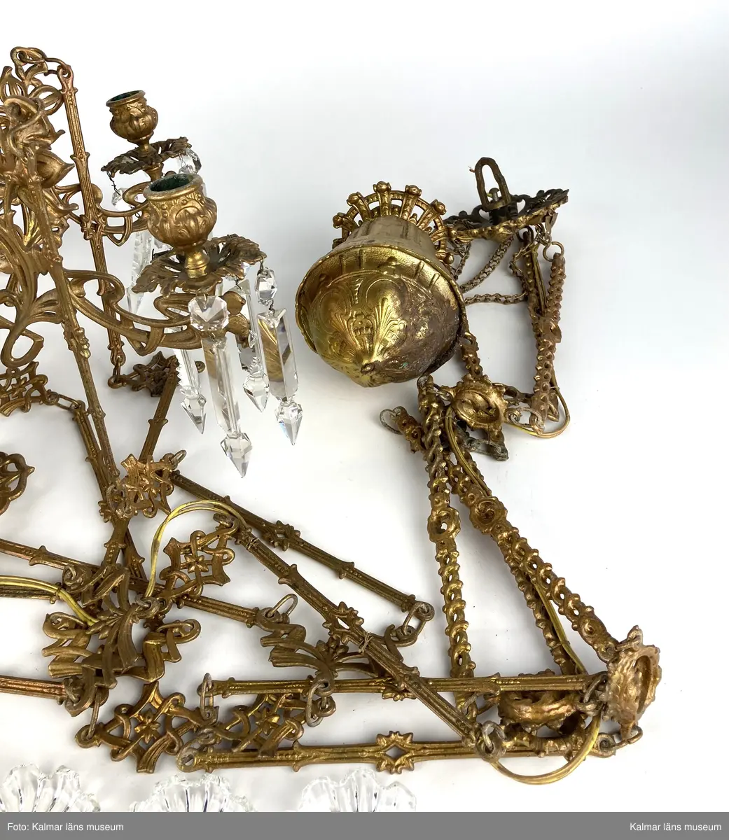 KLM 45619:390. Lampa, taklampa, fotogenlampa. Ombyggd fotogenlampa med stomme av bronserad gjutmetall samt keramik. Mittendelen är av glaserad keramik/porslin med gula blommor på stjälk med gröna blad. Bottenfärg i rödbrun nyans. Övre delen  är avtagbar med fastsatt glasbehållare undertill för fotogen. Lampan är ombyggd med dragen el och hållare för glödlampa. Sammanlagt 6 armar med hållare för stearinljus. Ljusarmarna är i snirklig jugendstil och är förgyllda. Ytterst på vardera arm hänger prismor av typen fotogenprisma. 6 tillhörande ljusmanschetter i genomskinligt glas. Glaskupa som sitter överst på stommen, i frostat vitt glas med sparade ytor som bildar mönster. Takkronan hänger i avlånga metalldelar som är ihopsatta med metallringar och fungerar på så vis som en kedja. Alla dekorerade med jugendmönster. Överst en hissanordning.