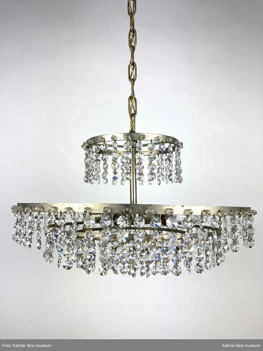 KLM 45619:392. Lampa, taklampa, kristallkrona. Av vit metall med fyra ringar av olika diametrar som hänger på olika höjd. På ringarna är prismor av sorten koppen fastsatta. På de två största ringarna sitter även pärlor fästa direkt i ringen. Eleiktrifierad med plats för 6 glödlampor.