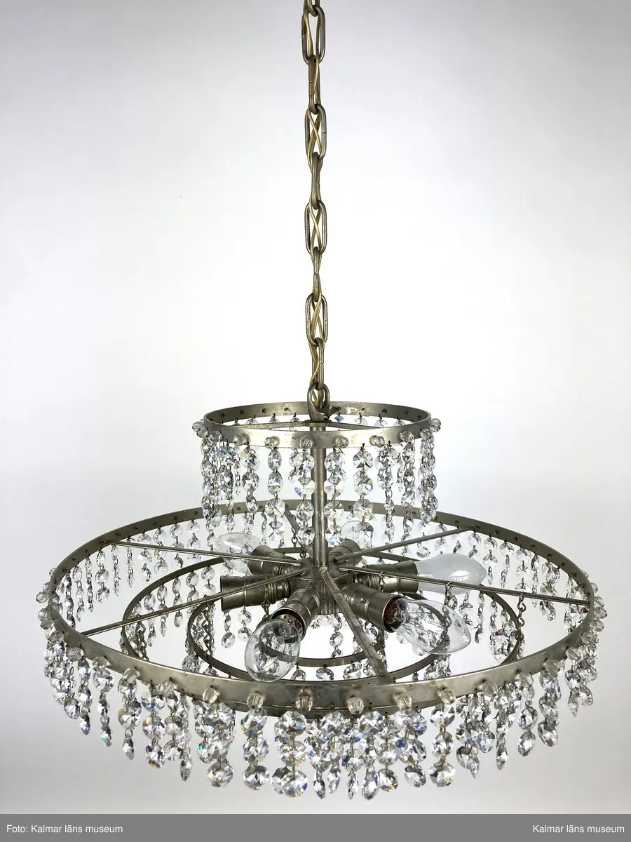 KLM 45619:392. Lampa, taklampa, kristallkrona. Av vit metall med fyra ringar av olika diametrar som hänger på olika höjd. På ringarna är prismor av sorten koppen fastsatta. På de två största ringarna sitter även pärlor fästa direkt i ringen. Eleiktrifierad med plats för 6 glödlampor.