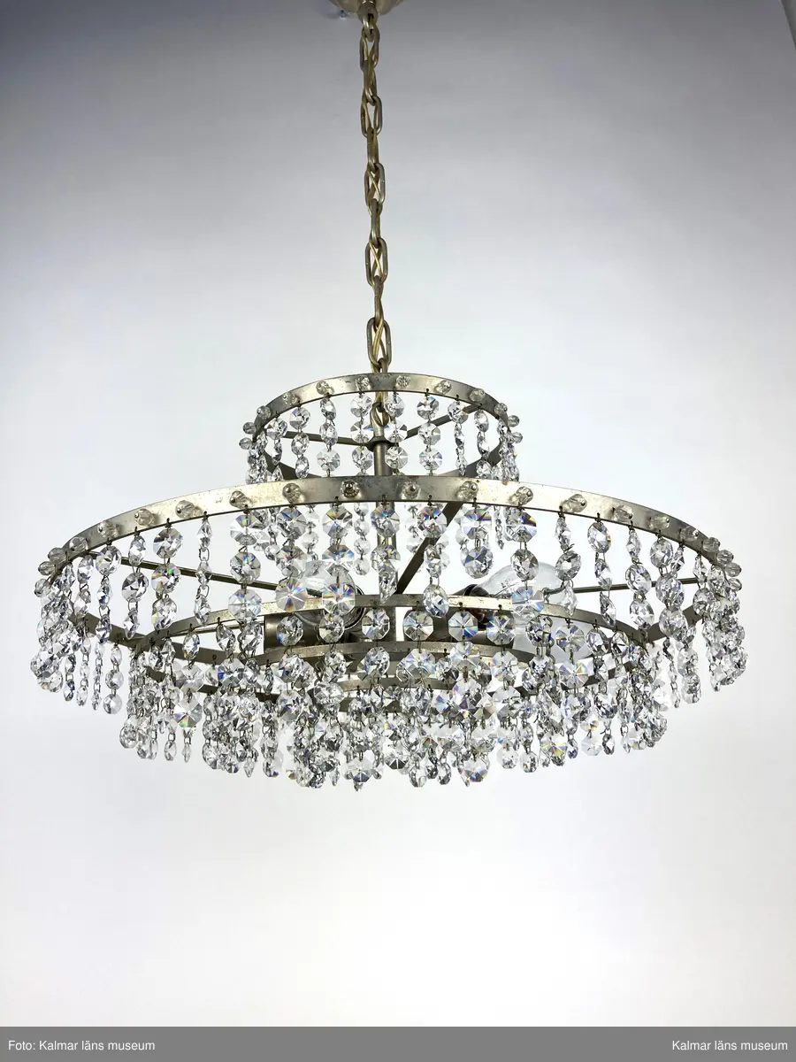 KLM 45619:392. Lampa, taklampa, kristallkrona. Av vit metall med fyra ringar av olika diametrar som hänger på olika höjd. På ringarna är prismor av sorten koppen fastsatta. På de två största ringarna sitter även pärlor fästa direkt i ringen. Eleiktrifierad med plats för 6 glödlampor.