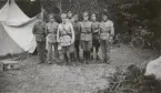 Åtta män i uniform m/1910 och m/1923 uppställda för fotografering utanför tält.