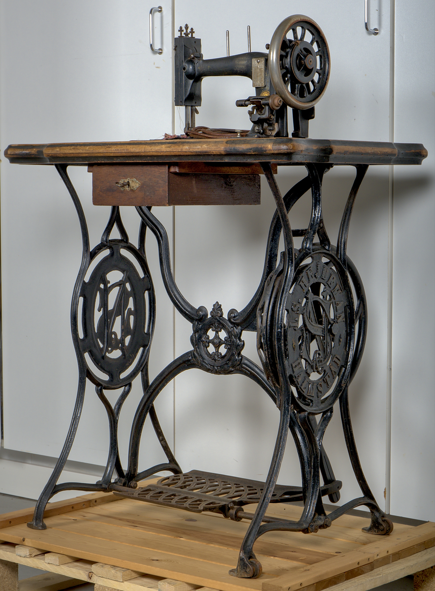 Trampsymaskin med dekorativt gjutjärnsunderrede och en träskiva som arbetsyta.

Svartmålad maskin dekorerad med sirlig ornamentik i guldfärg. På maskinens framsida sitter två märkta plåtar:

1. En referenstabell, för nålar?
2. Signering: "J.S. Hejdeman, Halmstad. Symaskin försäljning".

Maskinen är monterad på en profilerad träskiva av ek, försedd med en måttstock i intarsia på skivans framsida. Under bordet finns en skjutbar tillbehörslåda med låsmekanism. Maskinen skyddas av en rektangulär huv av ek, försedd med fyra dekorativa, svarvade hörnstolpar.

Det svartlackerade gjutjärnsstativet har en dekorativ utformning. På var sida av stativet finns två ovala medaljonger med relieftexten: "J.S. HEJDEMAN, HALMSTAD". De båda benen på höger sida av ställningen är försedda med hjul.