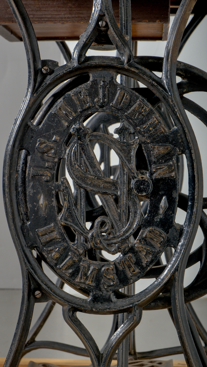 Trampsymaskin med dekorativt gjutjärnsunderrede och en träskiva som arbetsyta.

Svartmålad maskin dekorerad med sirlig ornamentik i guldfärg. På maskinens framsida sitter två märkta plåtar:

1. En referenstabell, för nålar?
2. Signering: "J.S. Hejdeman, Halmstad. Symaskin försäljning".

Maskinen är monterad på en profilerad träskiva av ek, försedd med en måttstock i intarsia på skivans framsida. Under bordet finns en skjutbar tillbehörslåda med låsmekanism. Maskinen skyddas av en rektangulär huv av ek, försedd med fyra dekorativa, svarvade hörnstolpar.

Det svartlackerade gjutjärnsstativet har en dekorativ utformning. På var sida av stativet finns två ovala medaljonger med relieftexten: "J.S. HEJDEMAN, HALMSTAD". De båda benen på höger sida av ställningen är försedda med hjul.