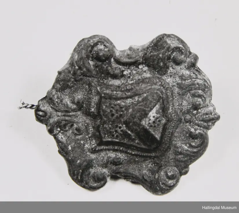 Form som byvåpen, støypt ornamentikk rundt (bladkrans). På baksida 4 naglar til å feste spennet.