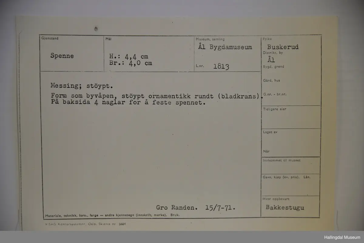 Form som byvåpen, støypt ornamentikk rundt (bladkrans). På baksida 4 naglar til å feste spennet.