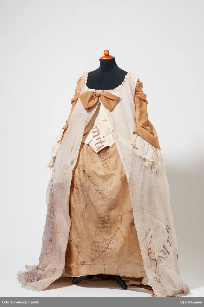 A= Overdel / snøreliv i silke sydd sammen med kappe 
i tyll med dekor i form av bokstaver
B= tynt sjal - to knyttet sammen
C= sjal i beige silkeaktig stoff dekorert med bokstaver
D= skjørt i beige og brunt silkeaktig stoff med dekor i form av bokstaver