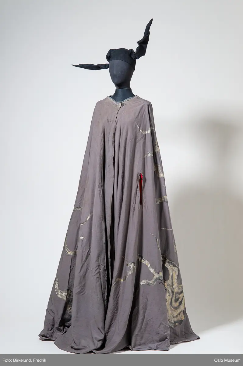 Lang cape i oransje stoff, med påmalte blomster og striper i grønt/blått/gull. Grått for.