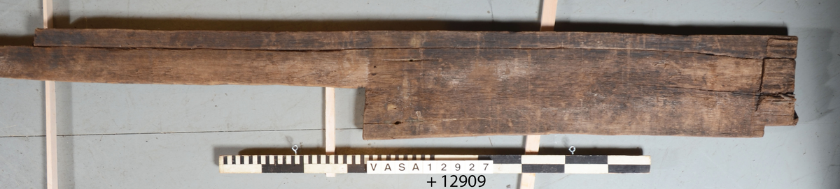 Del av planka. Nedtagen från skott i hålskeppet 2013. Plankan består av två delar, fnr 12909 och 12927. En bortspjälkad bit av den ena långsidan har vid något tillfälle återmonterats. Denna del har ett eget fyndnummer (12909). En kortare del av denna, en gång bortspjälkade kant, saknas fortfarande. Ett , med yxa, uthugget uttag för dörröppning finns mitt på plankan. Plankans ena ände är något fasad. Fem skruvhål varav ett är förstört av modern skruv.