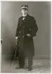 Nyblivne kronolänsmannen Ernst Bjersing, ca 1917, i länsmansuniform med rock och skärmmössa och med en portfölj under armen.
Helfigur, halvprofil. Ateljéfoto.