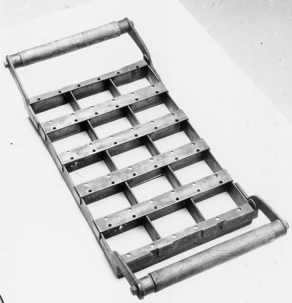 Rektangulär form av järn med två trähandtag längs kortsidorna. Formen är indelad i 15 lika stora rutor. Använd för bakning av skeppsskorpor så kallade knax vid Kronobageriet i Karlskrona.