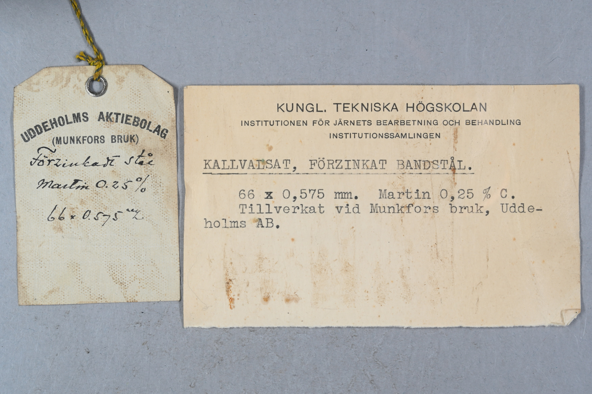 Prov på kallvalsat förzinkat bandstål. Två tillhörande lappar med notering: "Kallvalsat förzinkat bandstål. 66 x 0,575 mm. Martin 0,25 % C. Tillverkat vid Munkfors bruk, Uddeholms AB."