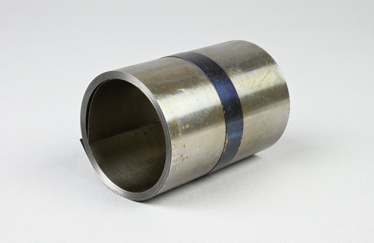 Prov på härdat bandstål. Två tillhörande lappar med notering: "Härdat bandstål. 89 x 0,15 mm. Martin 1,20 % C. Tillverkad vid Munkfors bruk, Uddeholms AB."