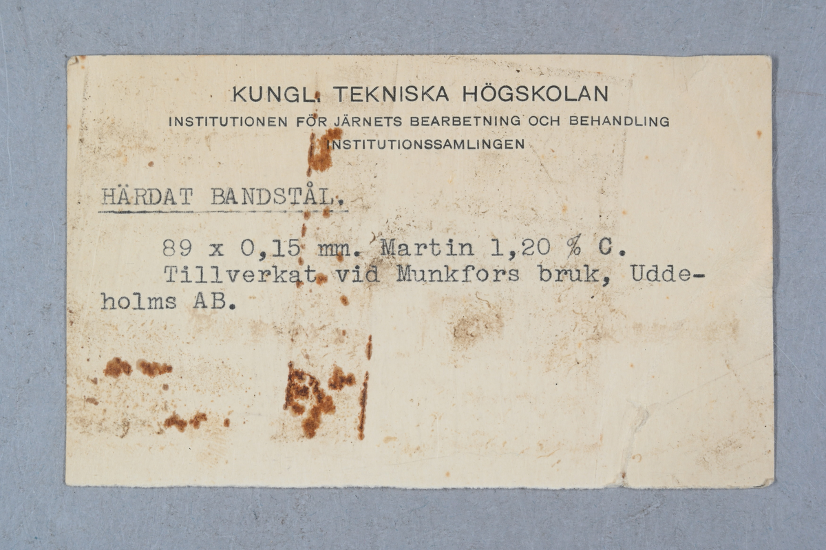 Prov på härdat bandstål. Två tillhörande lappar med notering: "Härdat bandstål. 89 x 0,15 mm. Martin 1,20 % C. Tillverkad vid Munkfors bruk, Uddeholms AB."