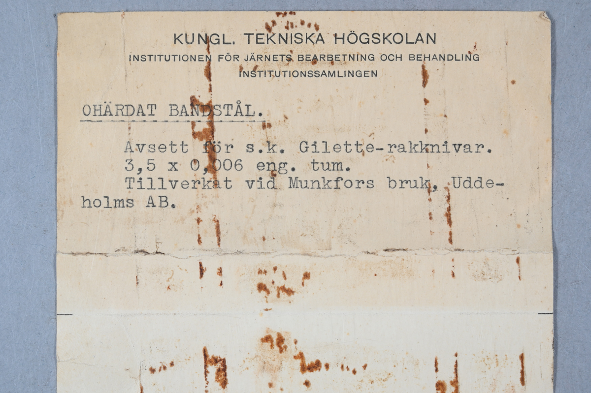 Prov på ohärdat bandstål. Två tillhörande lappar med notering: "Ohärdat bandstål. Avsett för s. k. Gilette-rakknivar. 3,5 x 0,006 eng. tum. Tillverkat vid Munkfors bruk, Uddeholms AB."