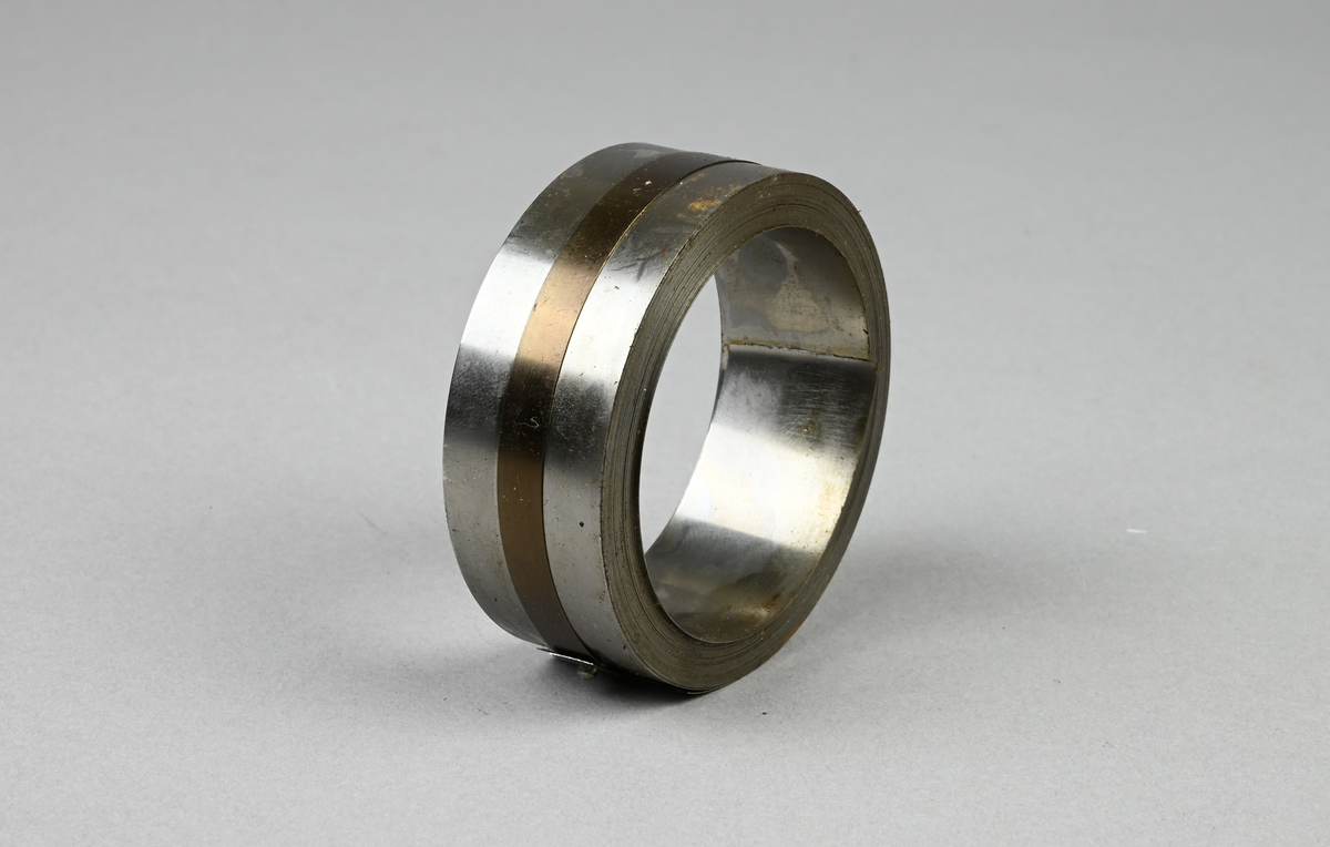 Prov på härdat bandstål. Två tillhörande lappar med notering: "Härdat bandstål för rakknivblad. 22,2 x 0,15 mm. Tillverkat vid Munkfors bruk, Uddeholms AB."