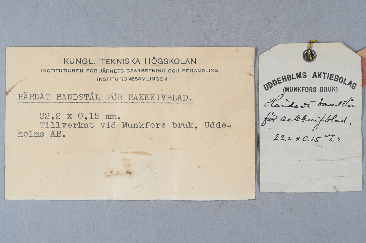 Prov på härdat bandstål. Två tillhörande lappar med notering: "Härdat bandstål för rakknivblad. 22,2 x 0,15 mm. Tillverkat vid Munkfors bruk, Uddeholms AB."
