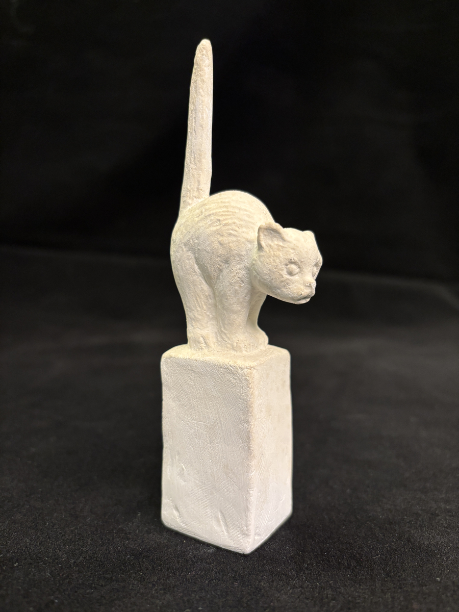 Gipsskulptur som föreställer en katt stående på en hög sockel.
Katten står med lätt böjd rygg och med svansen rakt upp.
På sockelns ena sida finns Carin Nilsons signering samt årtalet 1949.