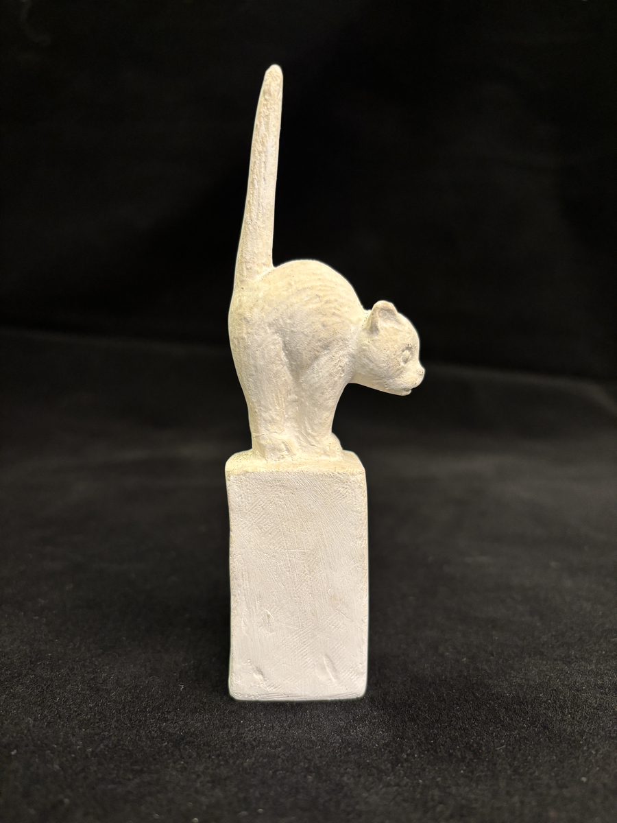 Gipsskulptur som föreställer en katt stående på en hög sockel.
Katten står med lätt böjd rygg och med svansen rakt upp.
På sockelns ena sida finns Carin Nilsons signering samt årtalet 1949.