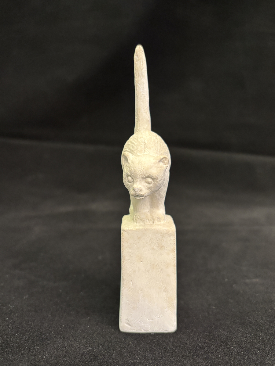 Gipsskulptur som föreställer en katt stående på en hög sockel.
Katten står med lätt böjd rygg och med svansen rakt upp.
På sockelns ena sida finns Carin Nilsons signering samt årtalet 1949.