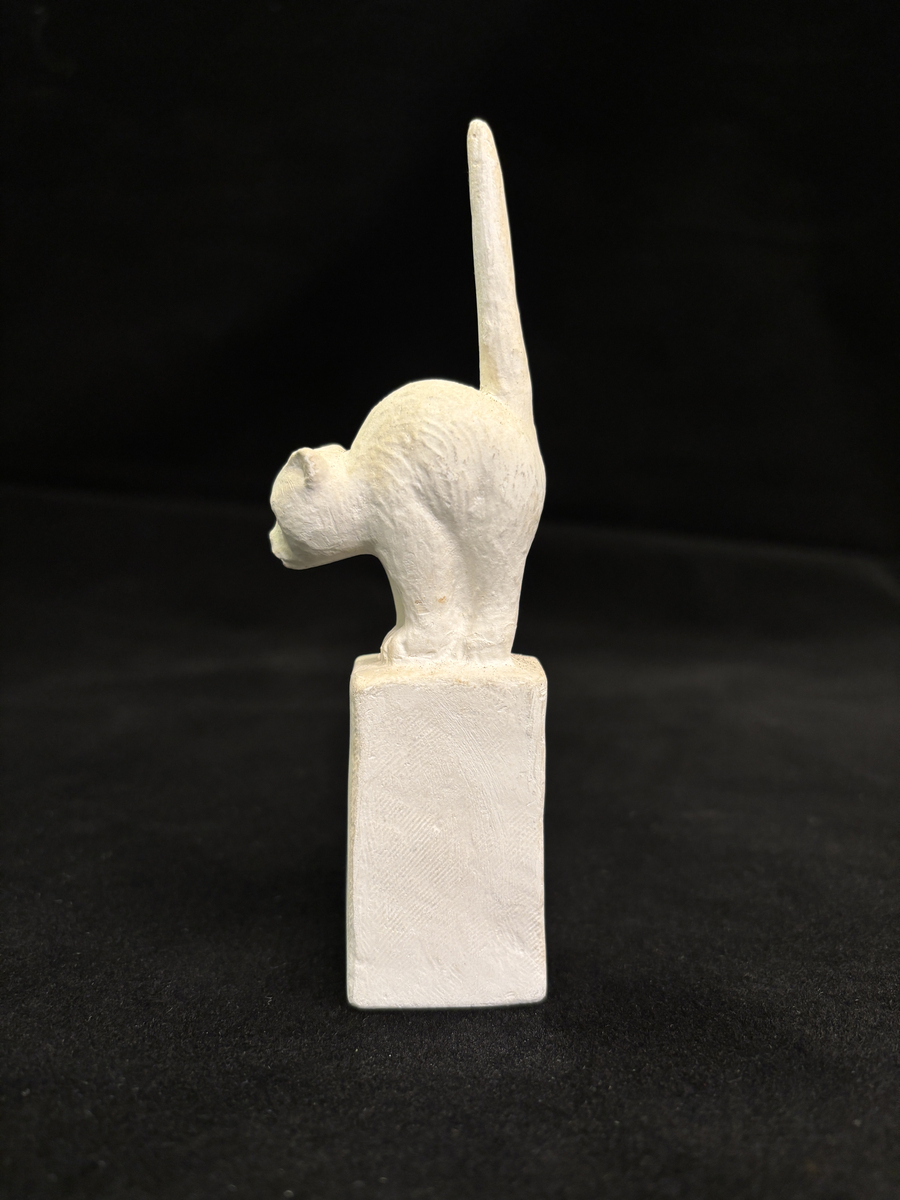 Gipsskulptur som föreställer en katt stående på en hög sockel.
Katten står med lätt böjd rygg och med svansen rakt upp.
På sockelns ena sida finns Carin Nilsons signering samt årtalet 1949.