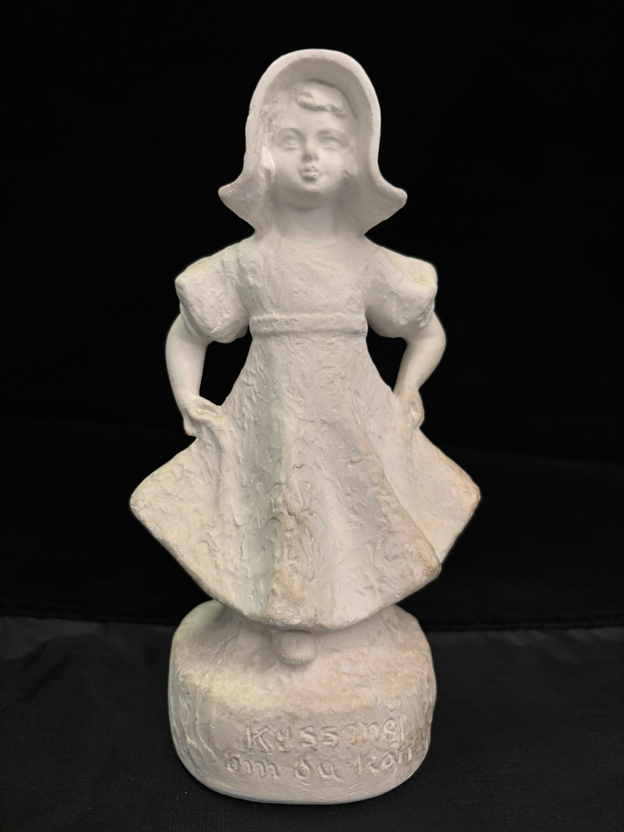 Gipsfigur. Skulptur som föreställer en flicka, stående på en sockel.
Flickan visas i helfigur. Hon är klädd i en klänning som hon håller i och ut med händerna och på huvudet har hon en hatt i bahyttstil, med stora brätten på sidorna.
På sockelns framsida står det "Kyss mej om du kan" och på andra sidan finns konstnärens signering: Carin Nilson samt årtal 1934.