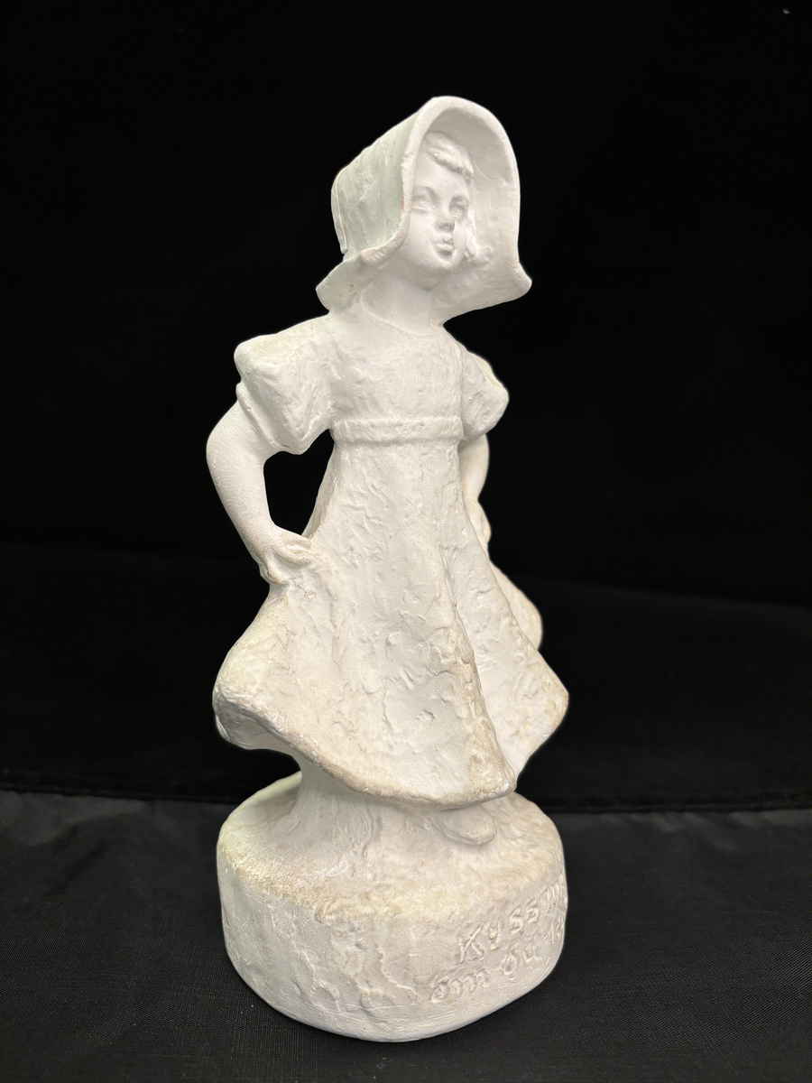 Gipsfigur. Skulptur som föreställer en flicka, stående på en sockel.
Flickan visas i helfigur. Hon är klädd i en klänning som hon håller i och ut med händerna och på huvudet har hon en hatt i bahyttstil, med stora brätten på sidorna.
På sockelns framsida står det "Kyss mej om du kan" och på andra sidan finns konstnärens signering: Carin Nilson samt årtal 1934.
