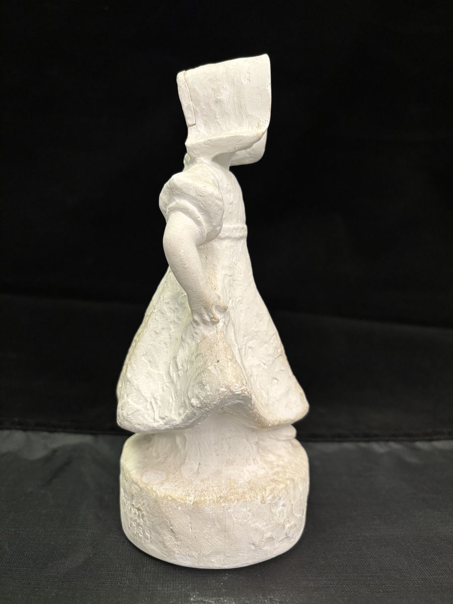 Gipsfigur. Skulptur som föreställer en flicka, stående på en sockel.
Flickan visas i helfigur. Hon är klädd i en klänning som hon håller i och ut med händerna och på huvudet har hon en hatt i bahyttstil, med stora brätten på sidorna.
På sockelns framsida står det "Kyss mej om du kan" och på andra sidan finns konstnärens signering: Carin Nilson samt årtal 1934.