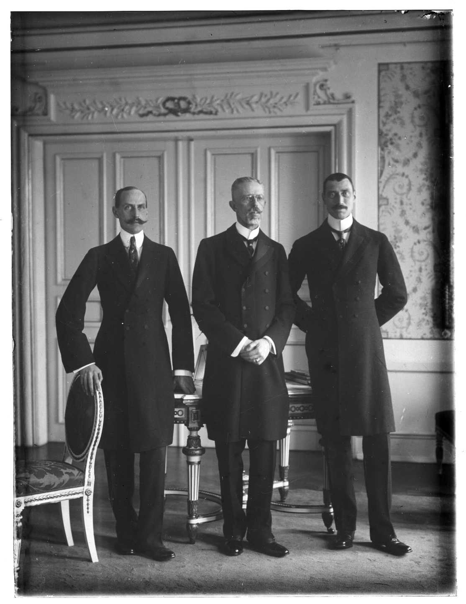 Tre menn i mørke jakker står side om side i et rom. Fra venstre mot høyre: Kong Haakon V av Norge, kong Gustav V av Sverige og kong Christian X av Danmark. I bakgrunnen er det en dobbel dør og mønstret tapet.