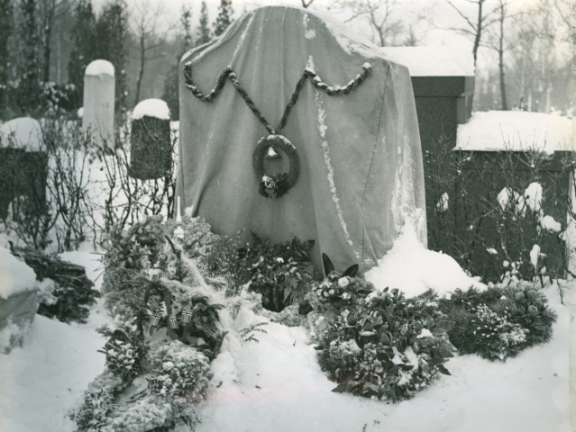 En gravstøtte som er tildekket og dekorert med julepynt. I bakgrunnen er det flere gravstøtter og trær, og bakken er dekket av snø.