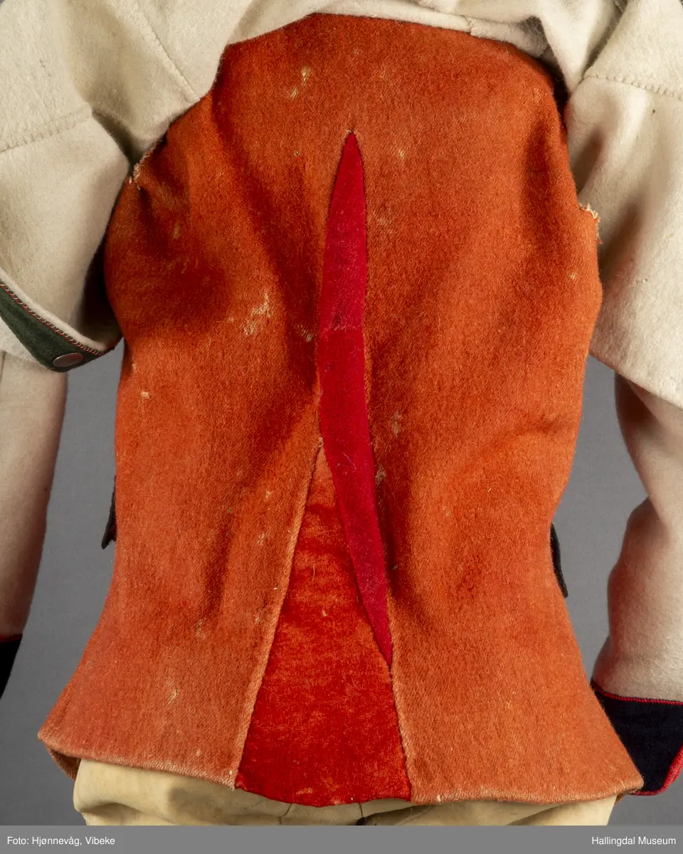 Vesten er sydd sammen av mange ulike røde stoffbiter. Ryggen av rødbrunt/orange vadmål. Nede på hver side i ryggen en liten kile av samme stoff, midt i ryggen felt inn en kile av rødbrun/orange stoff og 2 kiler av mørkt rødt stoff. Framstykkene og lommer er kantet med sort silkestoff. Framstykket og lommer er foret med lintøy.  Øverst på framstykket er foret med hvitt vadmål. 7 knapphull og 6 knapper på hver side framme. Danske mynter, III-skillinger fra 1727,1728,1730. To lommeklaffer med 3 knapphull i hver. Uten lommer. Knapphull sydd med grønn tynn 2-tråds ulltråd, Knappene er festet m/lærreim på innsiden. Nederst på hvert framstykke er bordert stilisert blomstermotiv i attersting og plattsøm av tynt 2-tråds ullgarn i gult, lys blått, grønt. Sydd og brodert for hånd.