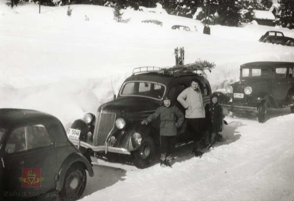 I midten: Ford V8 1936 Drosje med regnr. E-5046. Til høyre Fiat Balilla 1935 med regnr. D2982. I følge merking til bildet.
Ved siden av bilene står mor og to barn. Drosjen har ski på taket. Trolig påskeutfart i fint vintervær. 

Bilen til venstre er en Fiat 500 Topolino, en toseter kom først som 1936-modell, med motor på 569 cc og 13 hk. Topolino = Mikke Mus på italiensk. (Balilla, også kalt 508C og 1100), var en 4 dørs sedan med tilsvarende design, kom som 1938-modell.)
Opplysninger til kjøretøyene  fra Ivar Erlend Stav).