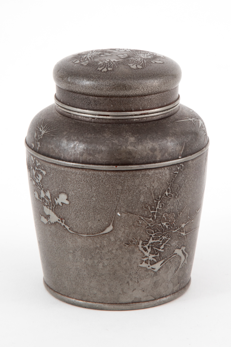 Støpt og dreiet vase i tinn. Rund form, med oppad litt utskrånende sider, avrundede skuldre og innsnevret hals. Dåsen har to lokk, det innerste med håndgrep. Overflaten er mørk ved etsing, og er dekorert med utsparret, strålignende mønster på korpus og lokk. Innsiden av korpus og det indre lokket er blankt.