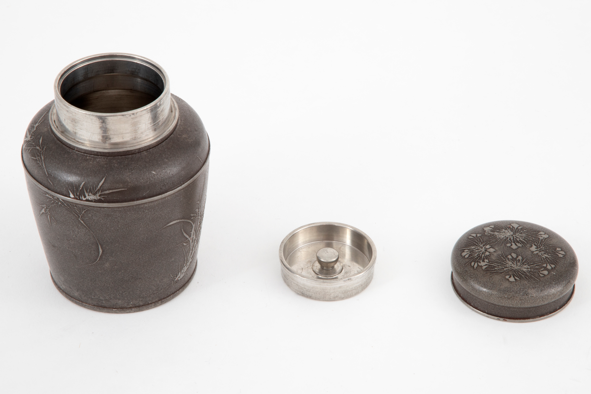 Støpt og dreiet vase i tinn. Rund form, med oppad litt utskrånende sider, avrundede skuldre og innsnevret hals. Dåsen har to lokk, det innerste med håndgrep. Overflaten er mørk ved etsing, og er dekorert med utsparret, strålignende mønster på korpus og lokk. Innsiden av korpus og det indre lokket er blankt.