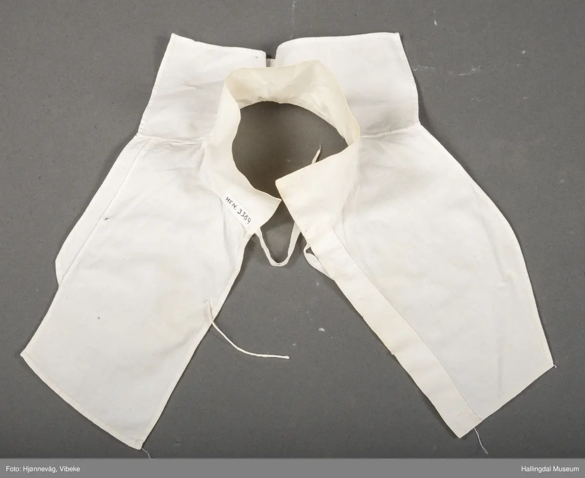 Bomullslerret hvitt håndsydd. Stående krage. Hold sammen framme med knyting av bendelbånd.