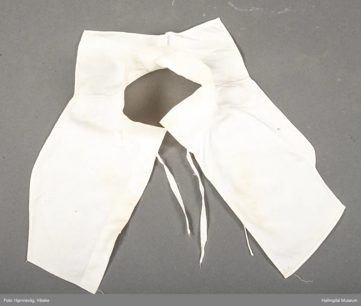 Bomullslerret hvitt håndsydd. Stående krage. Hold sammen framme med knyting av bendelbånd.